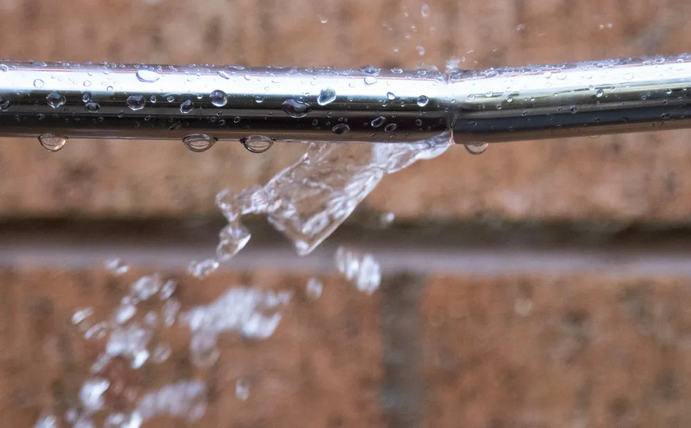Wasser schiesst aus einem Riss in einer Leitung vor einer Backsteinwand.